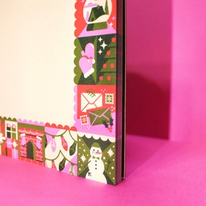Peut inclure: Un carnet d&eacute;coratif sur le th&egrave;me de No&euml;l. La bordure pr&eacute;sente des illustrations color&eacute;es d'une boule &agrave; neige, de moufles, d'enveloppes, d'un bonhomme de neige et d'autres &eacute;l&eacute;ments de vacances. Le carnet est pos&eacute; sur un fond rose vif.