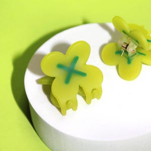 Pode incluir: Dois prendedores de cabelo verdes em forma de borboleta com um design de X verde. Os prendedores s&atilde;o feitos de pl&aacute;stico e t&ecirc;m um mecanismo de mola.