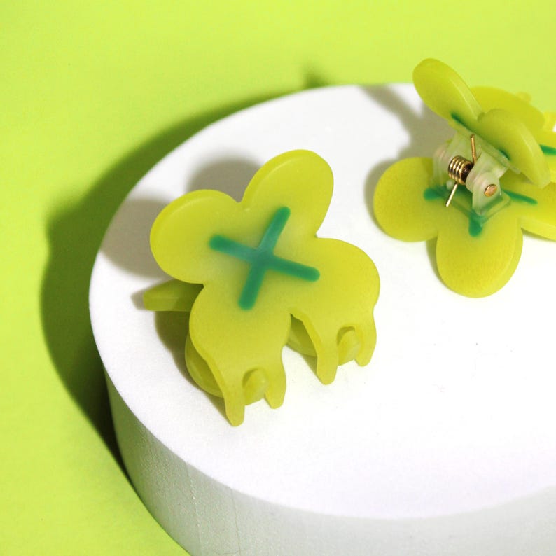 Pode incluir: Dois prendedores de cabelo verdes em forma de borboleta com um design de X verde. Os prendedores s&atilde;o feitos de pl&aacute;stico e t&ecirc;m um mecanismo de mola.