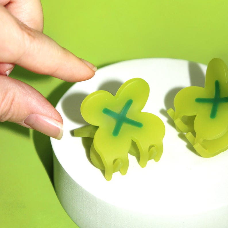 Pode incluir: Dois grampos de cabelo de pl&aacute;stico verde em forma de trevo de quatro folhas com um X verde no centro.