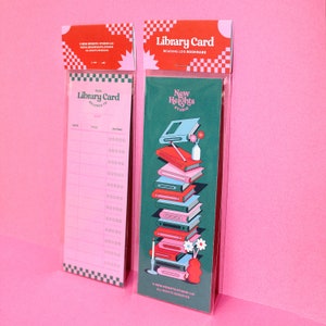 May include: A pink and green bookmark with a stack of books and the text "New Heights Studio" and "Library Card" on it.
