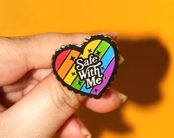 May include: A rainbow heart-shaped enamel pin with the text "Safe With Me" in black lettering.