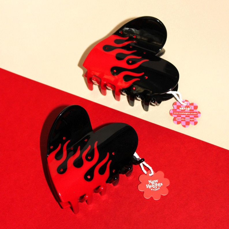 May include: Two heart-shaped hair clips with a flame design. The clips are black with red flames and have a claw clip closure. Each clip has a small tag attached with the text "hello!" and "New Heights Studio".