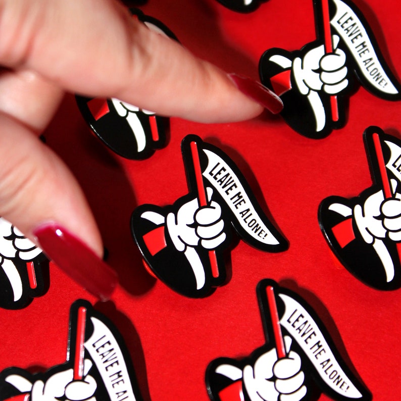 May include: A collection of black and red enamel pins with the text "Leave Me Alone!" on a red background.