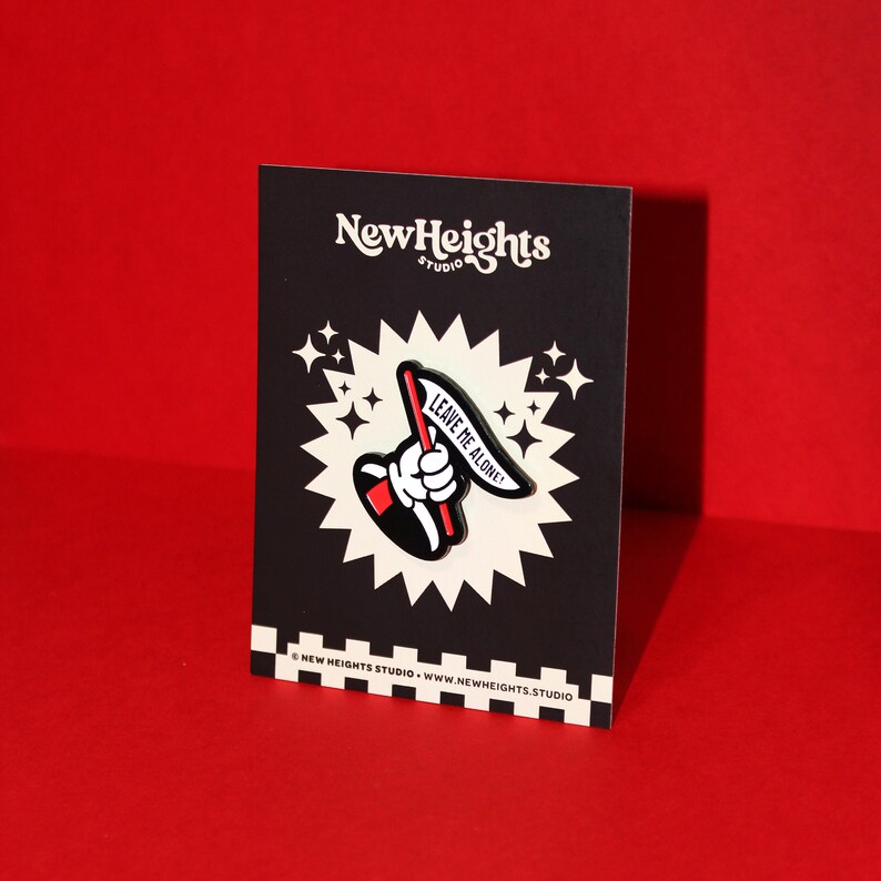 May include: Black and white enamel pin with a red and white flag that says "Leave Me Alone!" The pin is on a black card with white stars and the text "New Heights Studio" in white.