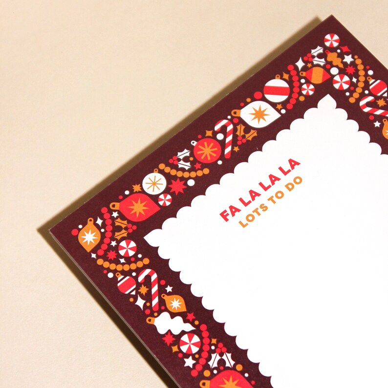 May include: A festive notepad with a dark red border adorned with Christmas ornaments, candy canes, and stars. The text "FA LA LA LA LOTS TO DO" is printed in red and orange. The notepad has a white, scalloped edge.