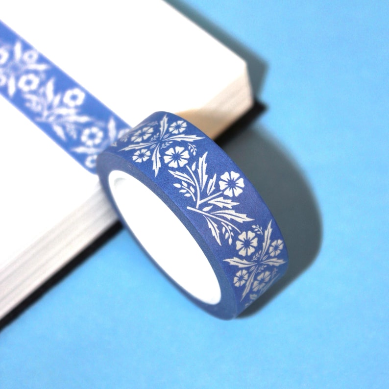 May include: A roll of blue washi tape with a white floral pattern. The tape is partially unrolled, revealing the design. The background is a light blue surface and a white notebook.