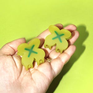 Pode incluir: Dois grampos de cabelo de pl&aacute;stico amarelo em forma de trevo de quatro folhas com um X verde no centro.