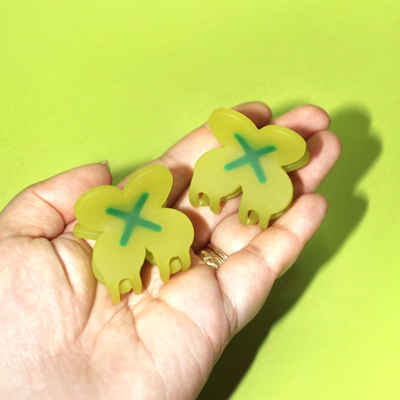 Pode incluir: Dois grampos de cabelo de pl&aacute;stico amarelo em forma de trevo de quatro folhas com um X verde no centro.