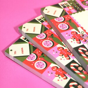 Peut inclure: Plusieurs piles d'&eacute;tiquettes cadeaux de No&euml;l avec un design festif. Les &eacute;tiquettes pr&eacute;sentent un motif rouge, rose et vert avec des bonhommes en pain d'&eacute;pices, des cannes en sucre et d'autres &eacute;l&eacute;ments de vacances. Chaque &eacute;tiquette a un espace pour &eacute;crire "&Agrave;:" et "DE:" sur un fond blanc.