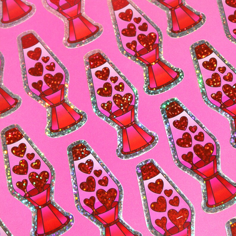 May include: A sheet of stickers featuring a lava lamp design filled with red glitter hearts. The lava lamps are pink and red with a silver glitter border, set against a bright pink background.