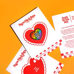 Op de afbeelding: Een witte kaart met een rood hartvormig ontwerp en de tekst "You are loved." De kaart ligt op een andere witte kaart met een rood hartvormig ontwerp en een regenboogkleurige emaille pin met de tekst "Safe With Me". De achtergrond is fel oranje.