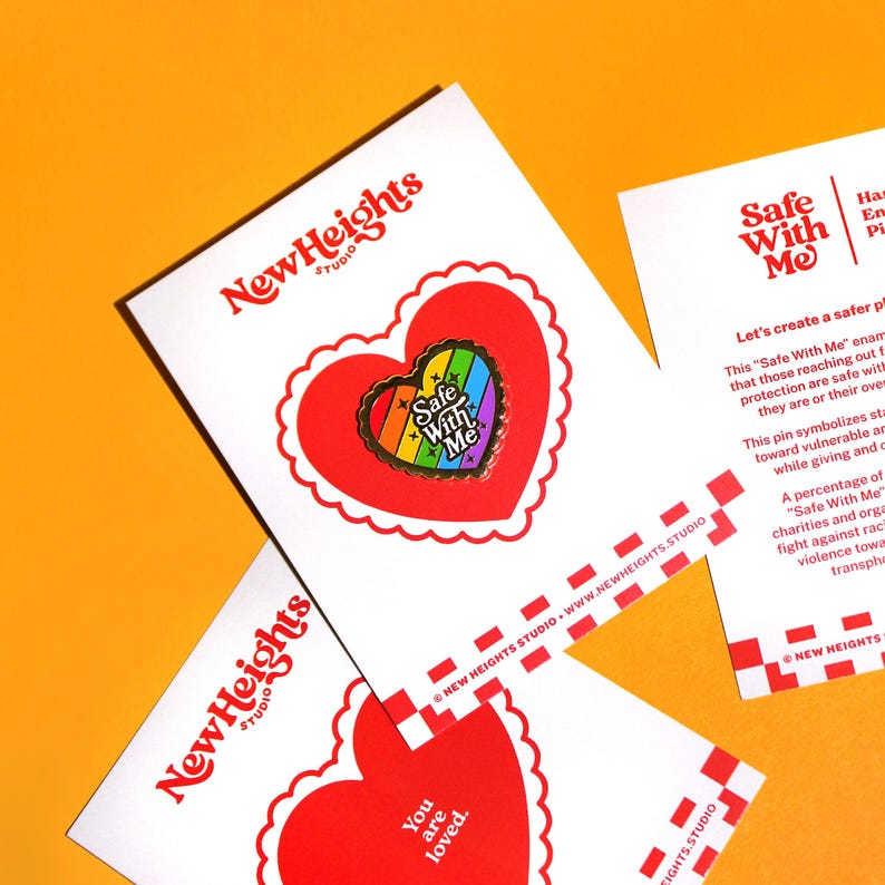 Op de afbeelding: Een witte kaart met een rood hartvormig ontwerp en de tekst "You are loved." De kaart ligt op een andere witte kaart met een rood hartvormig ontwerp en een regenboogkleurige emaille pin met de tekst "Safe With Me". De achtergrond is fel oranje.