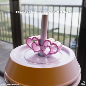 May include: A pink straw topper with a heart design, resting on a light pink lid. The straw is gray. The text "HEARTS STRAW TOPPER" is at the top of the image.