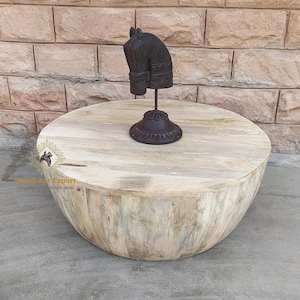 Puede incluir: Una mesa de centro redonda de madera con un acabado de madera clara. La mesa tiene una parte superior ligeramente curvada y una base ancha. Una escultura de cabeza de caballo de madera se coloca en la parte superior de la mesa.