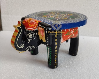 May include: Hand-painted wooden elephant stool with a round top. The elephant is black with colorful details, including red, yellow, green, and blue. The top of the stool is painted blue with a floral design.