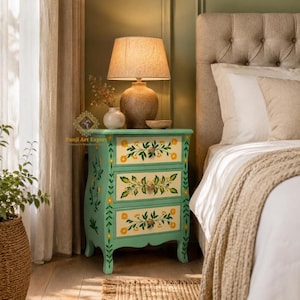 May include: A mint green bedside table with three drawers, adorned with yellow floral patterns and leaf accents. A beige lamp with a textured base sits atop the table, next to a vase and a small bowl. The table is placed next to a bed.