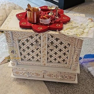 Puede incluir: Templo de madera blanco con tallas intrincadas y dos puertas. El templo tiene una bandeja de loto roja y blanca en la parte superior con una peque&ntilde;a olla de cobre, un peque&ntilde;o cuenco de plata y una cuerda roja y dorada.
