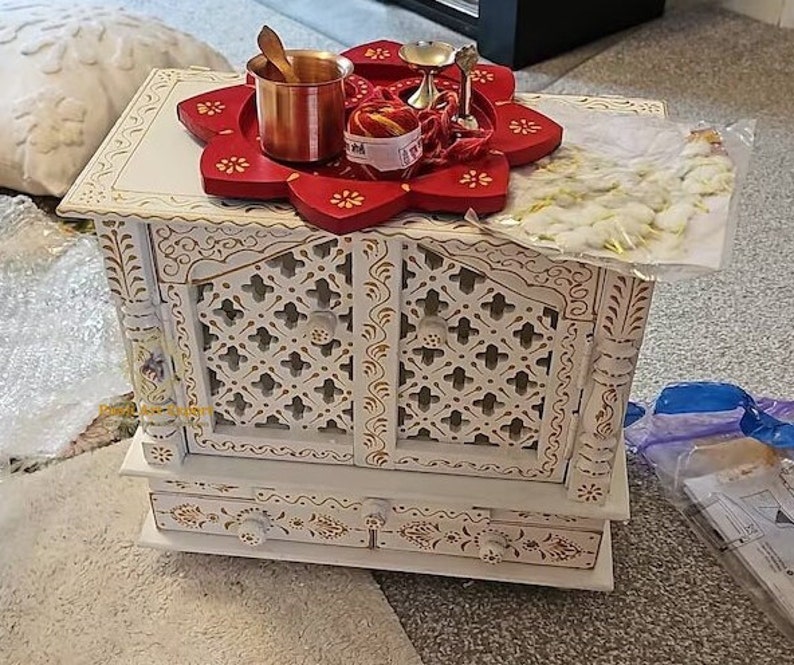 Puede incluir: Templo de madera blanco con tallas intrincadas y dos puertas. El templo tiene una bandeja de loto roja y blanca en la parte superior con una peque&ntilde;a olla de cobre, un peque&ntilde;o cuenco de plata y una cuerda roja y dorada.