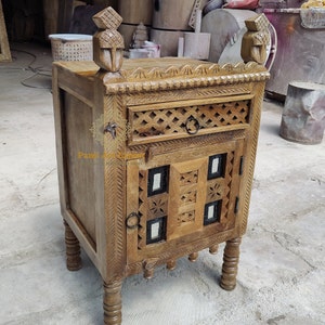 Op de afbeelding: Een houten bijzettafel met ingewikkelde houtsnijwerk en een enkele lade. De tafel heeft een donkerbruine afwerking en beschikt over een decoratieve deur met een tralieontwerp. De tafel wordt ondersteund door vier gedraaide poten.