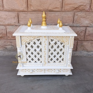 Puede incluir: Templo de madera blanco con detalles pintados en oro y una parte superior decorativa. El templo tiene dos puertas con un dise&ntilde;o de celos&iacute;a y un caj&oacute;n debajo. El templo est&aacute; sentado sobre una superficie gris.