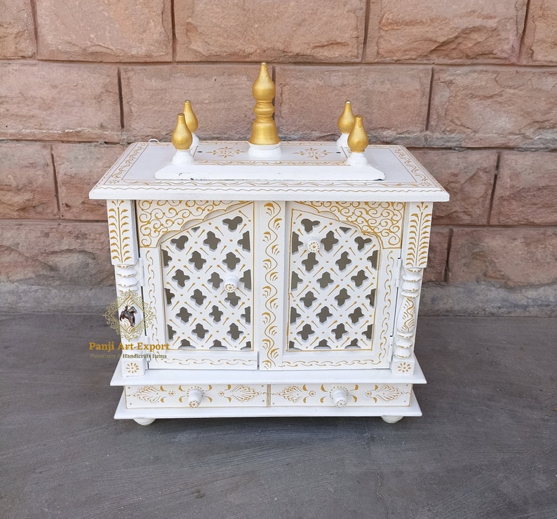 Puede incluir: Templo de madera blanco con detalles pintados en oro y una parte superior decorativa. El templo tiene dos puertas con un dise&ntilde;o de celos&iacute;a y un caj&oacute;n debajo. El templo est&aacute; sentado sobre una superficie gris.