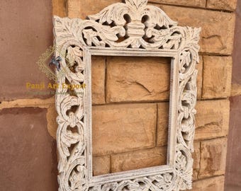 May include: Ornate, rectangular, off-white wooden frame with intricate floral carvings. The frame is mounted on a textured, light brown brick wall. The frame's design includes detailed leaf and scroll patterns.