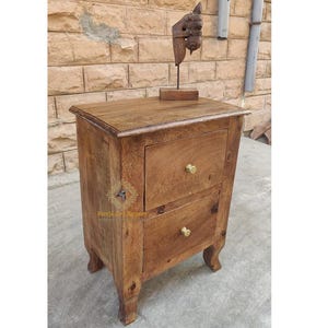 May include: A two-drawer wooden nightstand with brass knobs. The nightstand has a dark brown finish and sits on four legs. A wooden sculpture of a horse's head is on top of the nightstand.