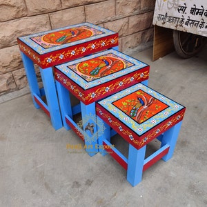 May include: A set of three nesting painted wooden stools. The stools are painted in shades of blue, red, and yellow with a peacock design. The largest stool is 16 inches wide, the middle stool is 12 inches wide, and the smallest stool is 8 inches wide.