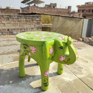 Puede incluir: Un taburete de elefante de madera verde con diseños de flores de loto rosas pintados en él. El taburete tiene una parte superior redonda y cuatro patas.