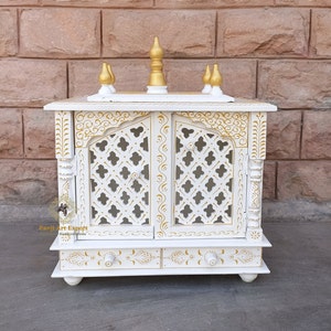 Puede incluir: Templo de madera blanco con detalles dorados y tallas intrincadas. El templo tiene dos puertas con un dise&ntilde;o de celos&iacute;a y un caj&oacute;n debajo. El templo est&aacute; decorado con pintura dorada y tiene un dise&ntilde;o tradicional indio.