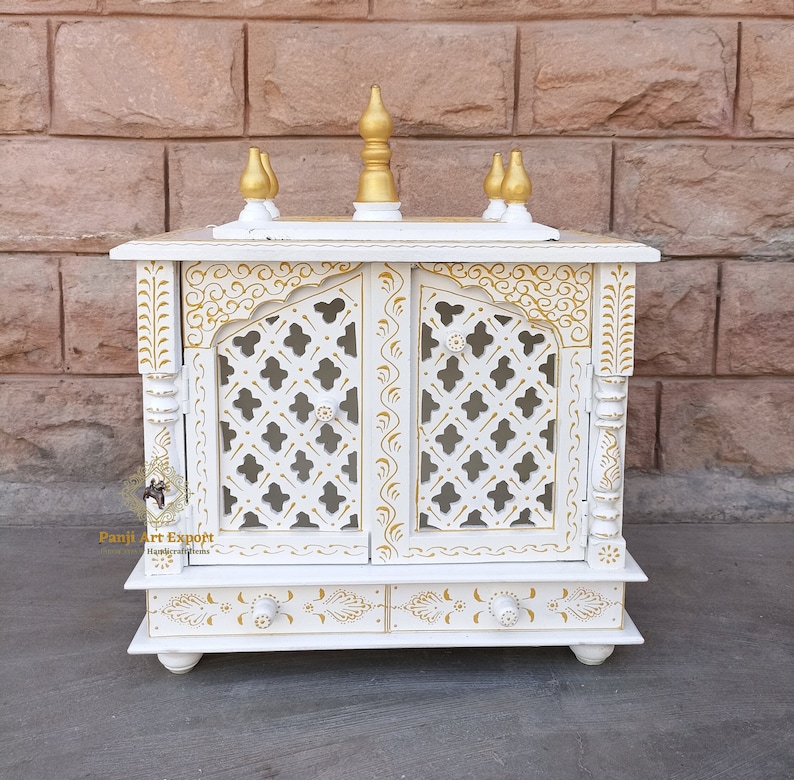 Puede incluir: Templo de madera blanco con detalles dorados y tallas intrincadas. El templo tiene dos puertas con un dise&ntilde;o de celos&iacute;a y un caj&oacute;n debajo. El templo est&aacute; decorado con pintura dorada y tiene un dise&ntilde;o tradicional indio.