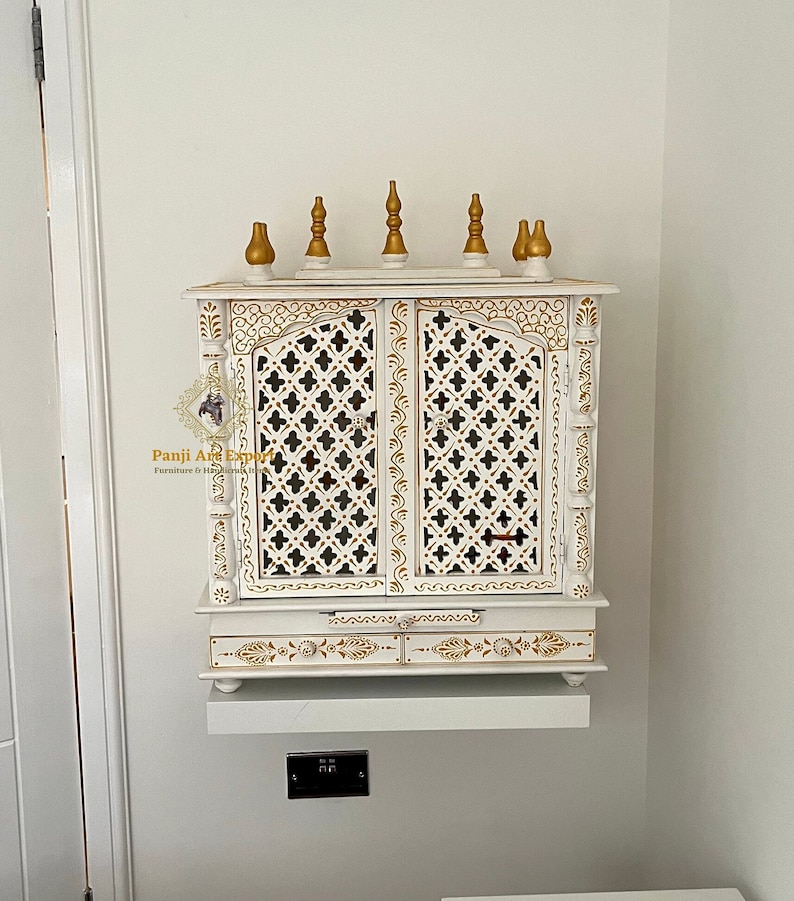 Puede incluir: Armario de madera blanco con detalles dorados y tallas intrincadas. El armario tiene dos puertas con un dise&ntilde;o de celos&iacute;a y un estante debajo. El armario est&aacute; montado en una pared y tiene un peque&ntilde;o estante debajo.