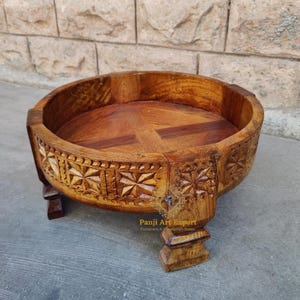 May include: A round wooden tray with intricate carvings on the sides and a base with four legs. The tray is made of light brown wood and has a natural finish.