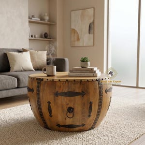 May include: A round wooden coffee table with metal accents. The table has a light brown finish and is decorated with dark metal bands and hardware. A mug and books sit on top of the table, which is placed on a neutral rug.