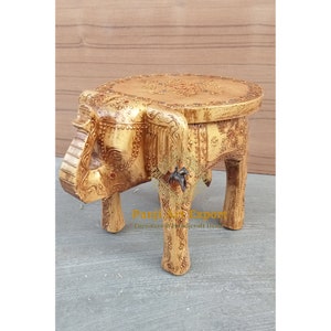 Op de afbeelding: Een handgesneden houten olifanten kruk met een ronde bovenkant en ingewikkelde houtsnijwerk. De kruk is geschilderd in een lichtbruine kleur met gouden accenten. De bovenkant van de kruk heeft een cirkelvormig ontwerp met een bloemmotief.