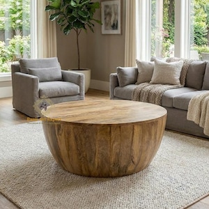 May include: A round, wooden coffee table with a natural finish sits on a light-colored rug in a living room setting. The table is surrounded by gray upholstered furniture and large windows.