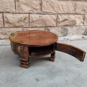 May include: A round wooden table with a hinged top that opens to reveal storage space. The table has carved details and sits on four legs.