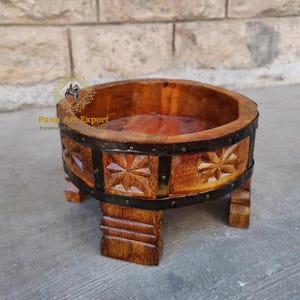 May include: A handcrafted wooden bowl with a dark brown finish, featuring carved starburst designs and metal accents. The bowl is supported by four short, sturdy legs. The product is from Panji Art Export.