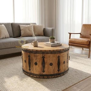 May include: A round coffee table made of light brown wood with dark metal detailing. The table has a drum shape with metal hardware. A mug, books, and a small plant are on the table.