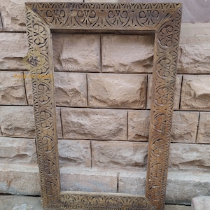 Puede incluir: Un marco de imagen de madera grande y tallado intrincadamente con un acabado marrón. El marco tiene una forma rectangular y presenta un borde decorativo con patrones florales y geométricos.