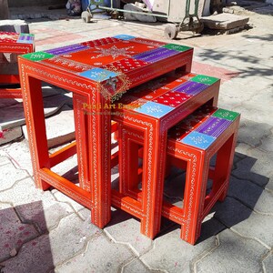 Puede incluir: Conjunto de tres mesas nido con intrincados diseños pintados a mano. Las mesas están hechas de madera y pintadas de rojo brillante con acentos coloridos. La mesa superior tiene un diseño de estrella en el centro.