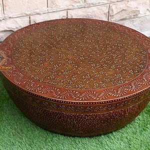 Pode incluir: Uma mesa de centro redonda de madeira castanho com padrões florais intrincados esculpidos na superfície. A mesa tem uma borda ligeiramente arredondada e um acabamento castanho escuro.