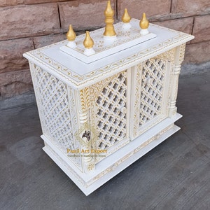 Puede incluir: Templo de madera blanco con intrincados tallados dorados y una parte superior decorativa con cinco remates dorados. El templo tiene una puerta con un dise&ntilde;o de celos&iacute;a y un caj&oacute;n debajo. El texto "Panji Art Export" es visible en la parte delantera del templo.