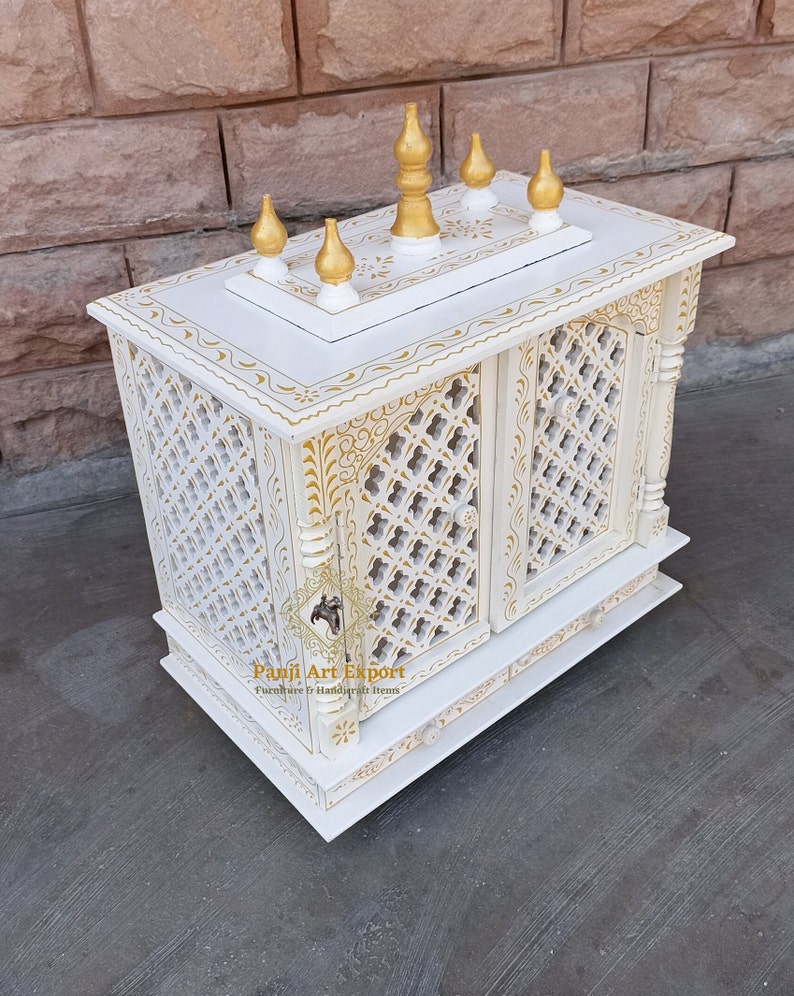 Puede incluir: Templo de madera blanco con intrincados tallados dorados y una parte superior decorativa con cinco remates dorados. El templo tiene una puerta con un dise&ntilde;o de celos&iacute;a y un caj&oacute;n debajo. El texto "Panji Art Export" es visible en la parte delantera del templo.