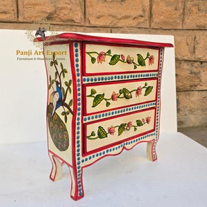 Puede incluir: Cómoda de tres cajones pintada a mano, con tapa y base rojas. Los cajones blanquecinos están decorados con motivos florales y detalles azules. Un pavo real está pintado en el lateral. Mueble único.