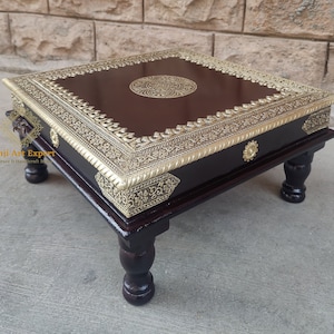 Pode incluir: Uma mesa quadrada de madeira castanho-escuro com quatro pernas torneadas. O tampo da mesa tem uma incrustação de latão decorativa com um desenho floral e um motivo circular no centro. A mesa tem aproximadamente 46 cm de largura.