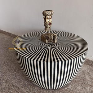 Puede incluir: Una mesa de centro redonda de incrustaciones de hueso con rayas blancas y negras, con una figurita decorativa en la parte superior. La mesa tiene un diseño único en forma de sol.