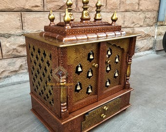 May include: A wooden temple with intricate carvings and gold accents. The temple has two doors with brass bells and a drawer at the bottom. The temple is decorated with a floral design.