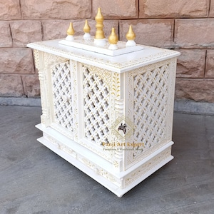 Puede incluir: Templo de madera blanco con intrincados tallados dorados y una parte superior decorativa con cinco remates dorados. El templo tiene dos puertas con un dise&ntilde;o de celos&iacute;a y un peque&ntilde;o caj&oacute;n debajo. El templo est&aacute; sentado sobre una superficie blanca.